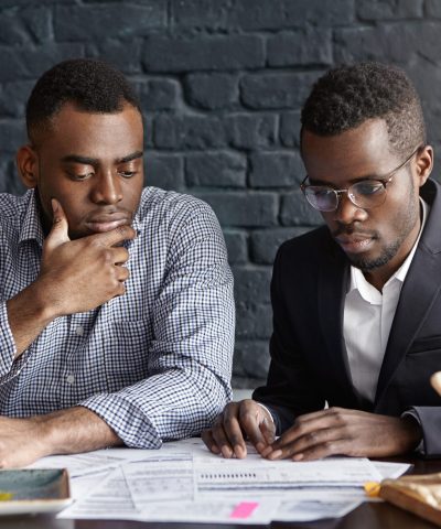 Two handsome dark-skinned executives having thoughtful and serious facial expression while reviewing finances and doing paperwork together, sitting at desk with documents in modern office interior